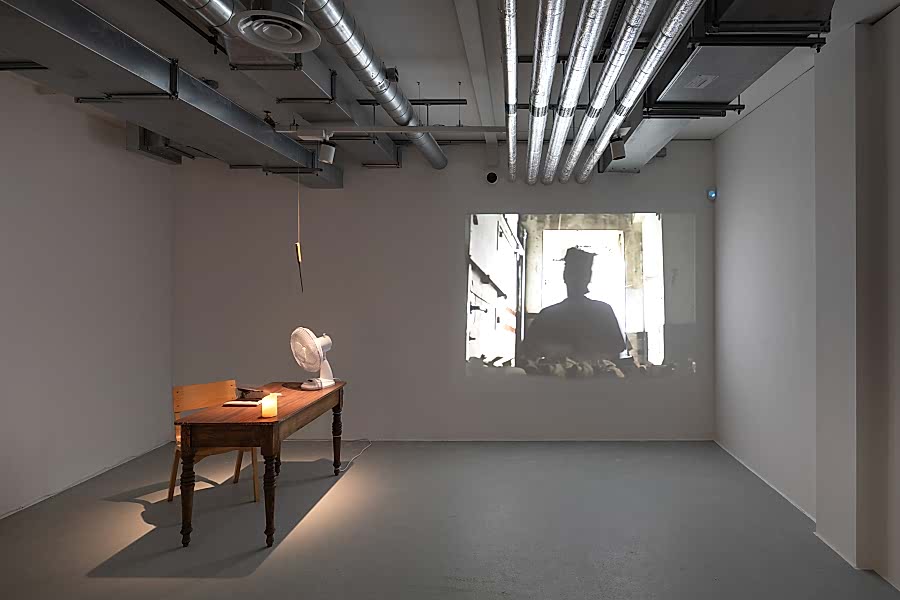Small dark gallery space with projected image on rear wall and a lit table with artworks